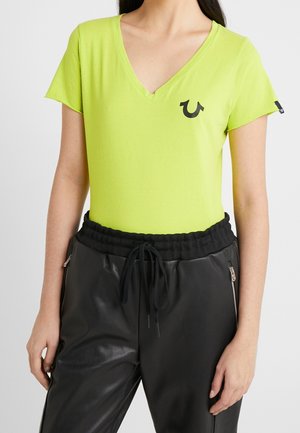 Kobieta w jasnym zielonym T-shircie z krótkim rękawem i dekoltem w serek, z czarnym logo na piersi, w zestawie z czarnymi spodniami na sznurek ze srebrnymi zamkami w kieszeniach.