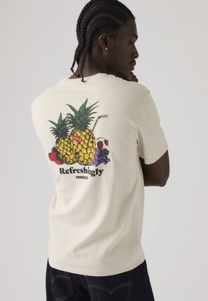 Homme aux cheveux tressés portant un t-shirt crème avec un ananas, des fraises, des raisins et le texte « Refreshingly smooth » au dos.