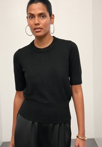 Maglione nero a maniche corte in maglia strutturata con orlo aderente, abbinato a una gonna di raso nera. Orecchini a cerchio grandi in oro completano il look.