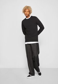 Zwarte sweatshirt met een ronde hals, gecombineerd met losse donkere broeken. De outfit heeft een wit shirt dat eronder is gelaagd. Zwarte sneakers maken de look compleet.