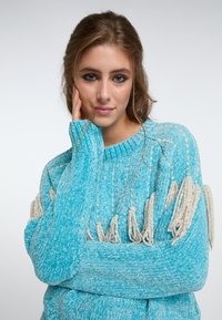 Maglione turchese lavorato a maglia con tessuto testurizzato, caratterizzato da dettagli in crema con frange sulle maniche e colletto a costine. Vestibilità morbida e oversize.