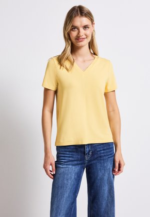 Żółty t-shirt z dekoltem w szpic i krótkimi rękawami, wykonany z gładkiego materiału, zestawiony z niebieskimi dżinsami o szerokich nogawkach. Prosty design, brak widocznych wzorów.