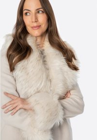 Płaszcz z sztucznego futra w jasnym beżu z puszystym białym kołnierzem i mankietami. Tkanina wydaje się miękka o gładkiej fakturze.