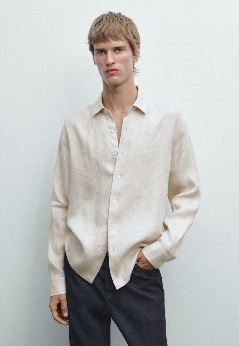 Linen button-up shirt in light beige with a relaxed fit, long sleeves, and a classic collar; paired with dark denim pants.