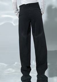 Inimene, kes kannab musta triibulist laiade säärtega pükse ja valget pikkade varrukatega särk, seisab taskutes kätega, seljaga vaataja poole.