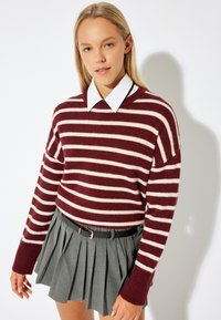 Maglione a righe color bordeaux e crema con una vestibilità comoda, abbinato a una gonna mini grigia plissettata e una cintura nera. Camicia bianca con colletto sotto.