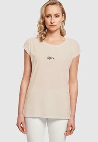 Beige bomull t-skjorte med rund hals og korte rullede ermer. Har svart brodert tekst "Inspire." på fronten.