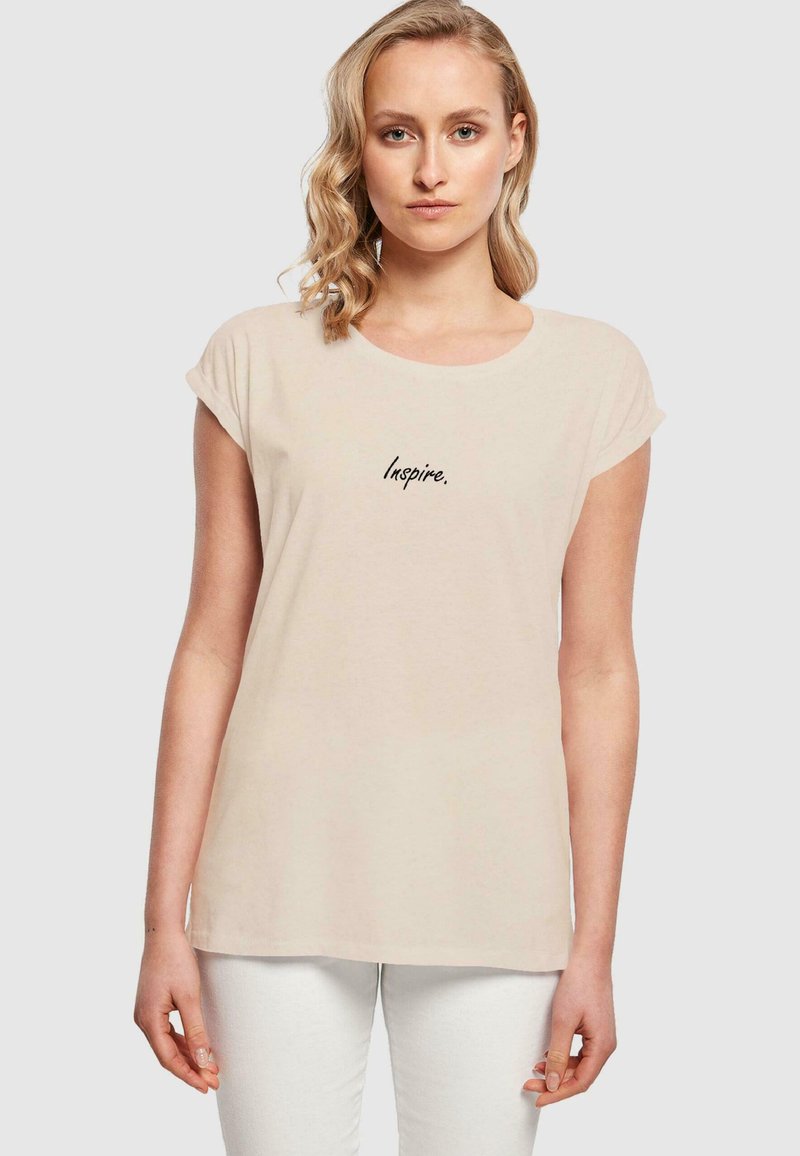 Beige bomull t-skjorte med rund hals og korte rullede ermer. Har svart brodert tekst "Inspire." på fronten.