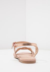 Vaaleanpunainen kultainen litteä sandaali, jossa on metallinen hihna, pehmeä vaaleanpunainen pohja ja avonainen muotoilu. Varustettu kantahellolla ja teksturoidulla hihna-aksentilla.