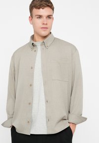 Μπεζ πουκάμισο με κουμπιά και τσέπη στο στήθος, φτιαγμένο από υφασμάτινη ύφανση, φορεμένο πάνω από γκρι μπλούζα. Διαθέτει γυρισμένα μανίκια και καφέ κουμπιά.