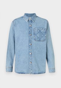 Camisa de denim azul claro com fecho de botões à frente, colarinho pontudo e um único bolso no peito com um padrão geométrico subtil.