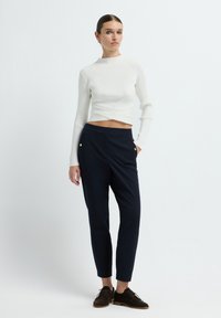 Witte geribbelde cropped trui met een twist-design, gecombineerd met marineblauwe tapered broeken met gouden knoopaccenten en een gladde textuur.