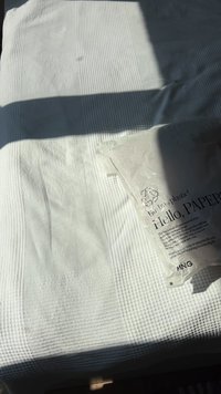 Bijela teksturirana tkanina prebačena preko površine, s nabranim i glatkim dijelovima. Vidljiva je zgužvana prozirna vrećica s oznakom "Hello, PAPER".