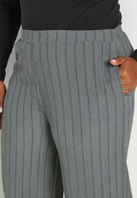 Grå ribbade byxor med bred midjeband, prydda med vertikala randmönster och sidofickor. Tyget har en mjuk, texturerad yta.