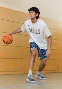 Tricou alb Nike cu imprimeu "BULLS", pantaloni scurți cu un model albastru, adidași gri cu talpă transparentă, dribling o minge de baschet portocalie pe un teren de baschet interior.