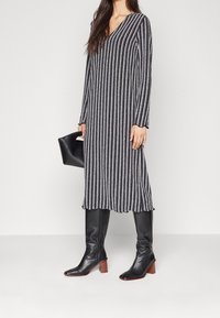 Robe tricotée à rayures verticales en bleu marine et gris, avec un col en V, des manches longues et un ourlet festonné. Associée à des bottes noires jusqu'aux genoux.