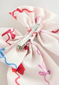 Un clip à cheveux avec un grand nœud rose pastel orné de motifs de rubans rouges et bleus. Une pince en métal argenté au centre. Tissu doux et texturé.