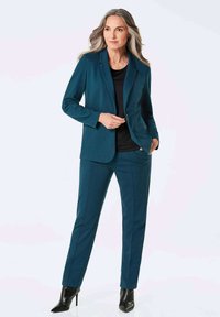 Teal blazer en bijpassende taperede broek van een gladde stof. De blazer heeft een enkele knoop en gekartelde revers; gecombineerd met zwarte hakken.
