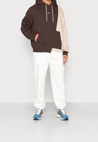 Felpa a blocchi di colore marrone e beige con tasca anteriore e cordini, abbinata a pantaloni della tuta bianchi e sneakers blu con motivo.