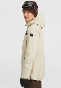 Beige jas met capuchon, zwarte accenten en ritszakken. Model draagt een bruine beanie en skibril, handen bedekt met zwarte handschoenen.