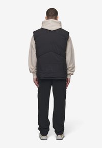 Czarna pikowana kamizelka z wysokim kołnierzem, noszona na beżowej bluzie z kapturem. Posiada tonalne logo z tyłu oraz spodnie cargo z kieszeniami.