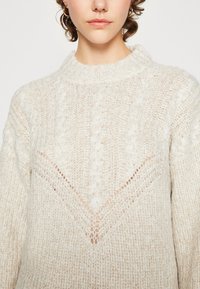 Cremefarvet strikket sweater med høj krave, der har et struktureret kabelstrikket mønster og en V-formet udskæring under halsudskæringen.