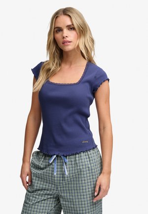 Femme blonde portant un haut bleu ajusté à manches courtes avec des bords festonnés et un pantalon à carreaux à cordon sur fond blanc.