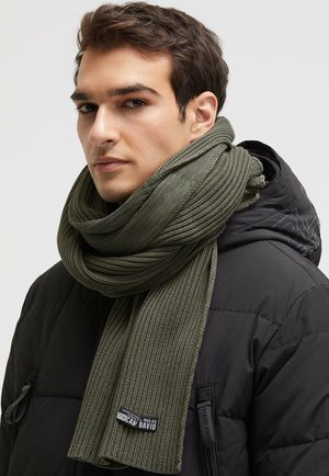 Olivgrüner, gerippter Strickschal, der um den Hals drapiert ist und ein Logoetikett trägt, kombiniert mit einer schwarzen Daunenjacke vor einem neutralen Hintergrund.