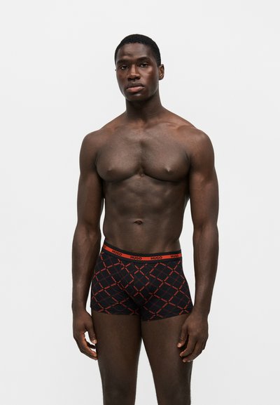 Homme athlétique à la peau foncée debout, portant un boxer noir avec un motif diagonal de logo rouge et une ceinture rouge sur fond blanc uni.