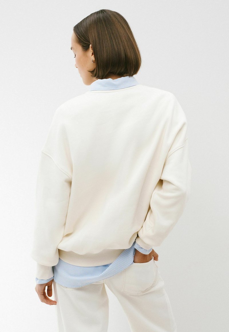 Personne aux cheveux courts et bruns portant un pull crème oversize par-dessus une chemise à rayures bleues et blanches et un pantalon crème, debout avec une main dans la poche.