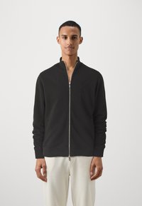 Juoda zip-up džemperis su aukštu apykakle, pagamintas iš minkštos medžiagos. Yra įrengti raukšlėti rankogaliai ir subtilus logotipas ant krūtinės. Dėvimi su šviesiomis kelnėmis.