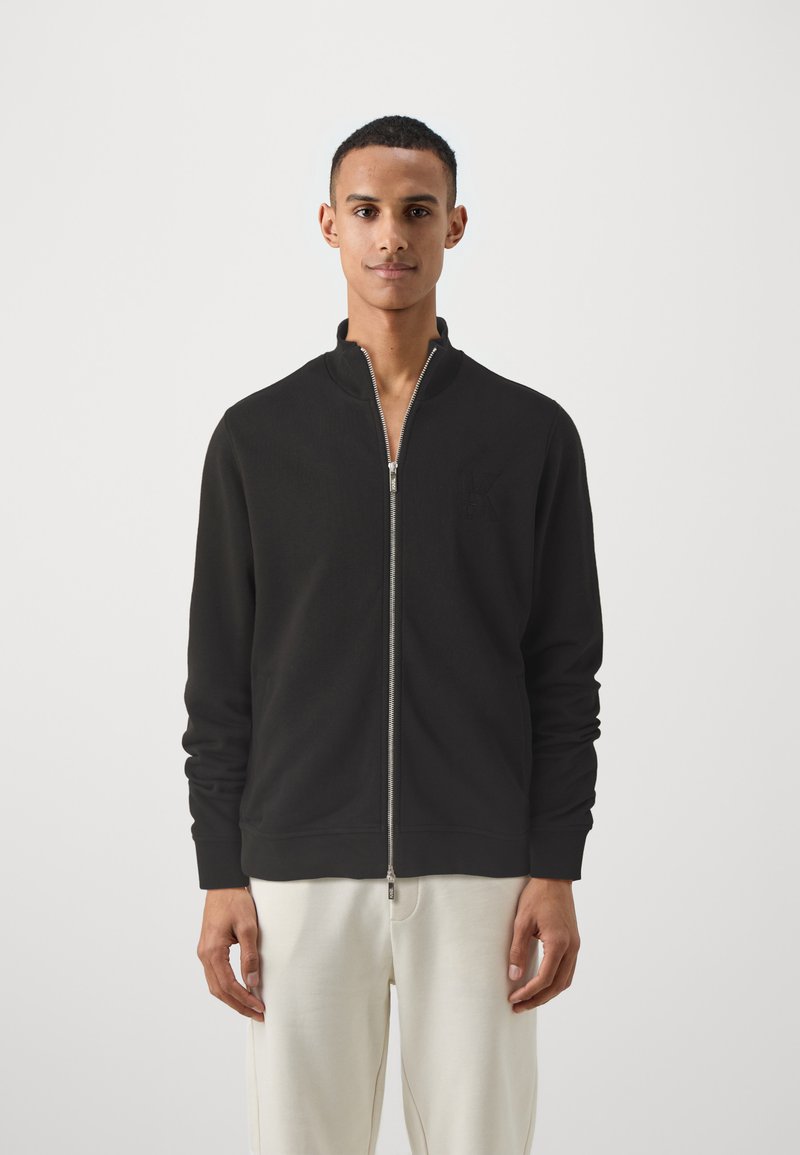 Juoda zip-up džemperis su aukštu apykakle, pagamintas iš minkštos medžiagos. Yra įrengti raukšlėti rankogaliai ir subtilus logotipas ant krūtinės. Dėvimi su šviesiomis kelnėmis.