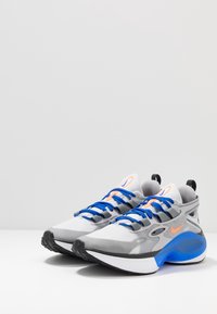 Graue Sportschuhe mit blauen Schnürsenkeln, orangefarbenen Akzenten und einer strukturierten Oberseite. Verfügt über eine schwarze Gummisohle und eine abgerundete, gepolsterte Sohle.