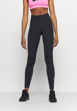 Mujer con mallas negras de tiro alto, zapatillas negras y un sujetador deportivo rosa de Nike frente a un fondo liso.
