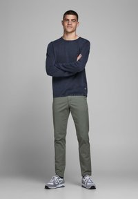 Mornarska majica z dolgimi rokavi, zelene hlače slim-fit in sivi športni čevlji z belimi poudarki. Model stoji s prekrižanimi rokami ob nevtralni ozadju.