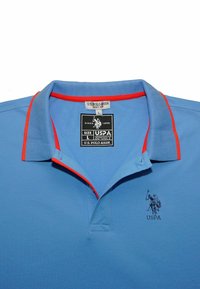 Modrá polo košeľa s červenými akcentmi na golieri, s logom na nášivke a textúrovanou tkaninou. Veľkostná nálepka: L, značka: U.S. Polo Assn.