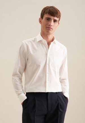 Jeune homme portant une chemise blanche boutonnée avec poche poitrine et un pantalon plissé noir, debout les mains dans les poches devant un fond uni.