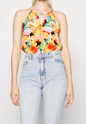 Kobieta w bezrękawniku w kwiaty w żółtym, pomarańczowym i różowym kolorze, w połączeniu z jasnoniebieskimi jeansami z wysokim stanem, na tle białym.