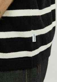 Stribet sort og hvid striktrøje med en blød tekstur og et lille mærke med teksten "JACK & JONES" på kanten. Løs, komfortabel pasform.
