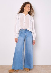 Weiße Bluse mit langen Ärmeln, gestickten Details und Knopfkragen, kombiniert mit hoch taillierten, hellblauen, weiten Jeans und braunen Clogs.