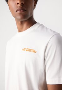 Weißes Baumwoll-T-Shirt mit orangefarbener bedruckter Aufschrift "Dein Abenteuer" und "Marc O'Polo Denim" auf der linken Brustseite. Kurze Ärmel, Rundhalsausschnitt.