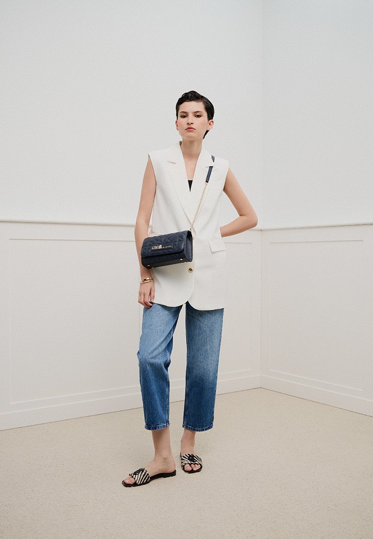 Mujer con cabello corto lleva un blazer blanco sin mangas, jeans azules, sandalias de patrón de cebra y lleva un bolso de hombro acolchado azul marino con cadena.