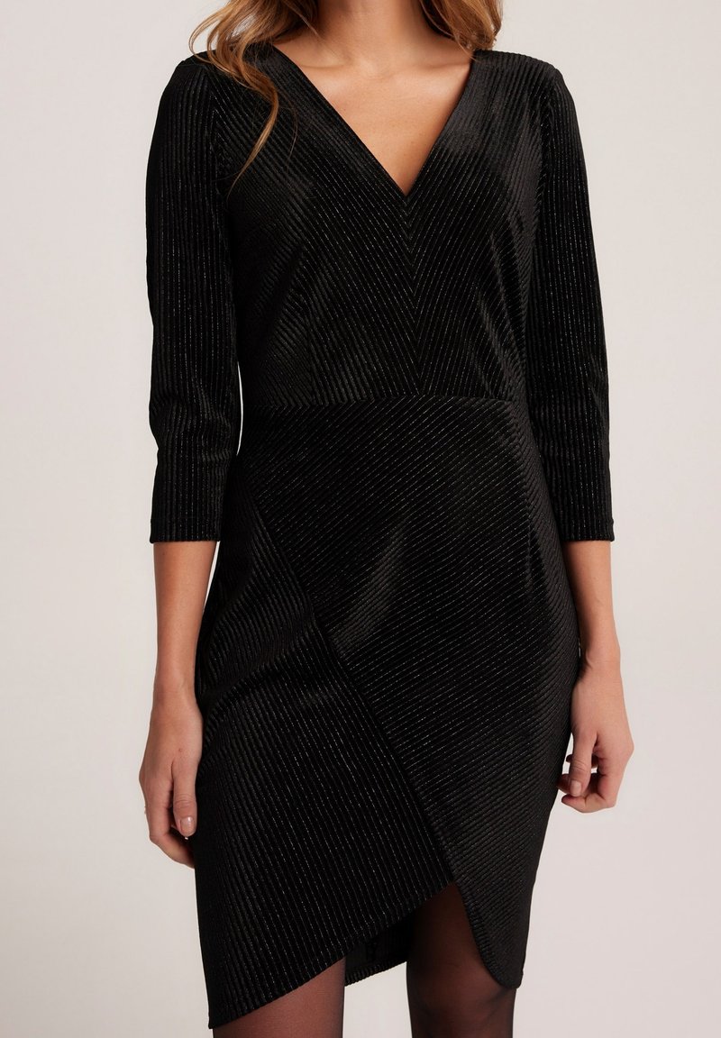 Robe en velours noir avec texture côtelée diagonale, décolleté en V, manches trois-quarts et détail asymétrique sur la jupe portée par une femme.