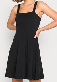 Svart ärmlös klänning med fyrkantig halsringning, figurnära liv och utsvängd kjol. Tillverkad av ett slätt tyg med en ren design och inga mönster.