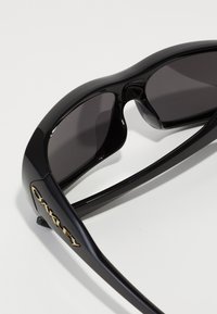 Óculos de sol Oakley pretos e elegantes com lentes escuras, exibidos dobrados sobre uma superfície branca, destacando a moldura brilhante e o logótipo dourado na haste.