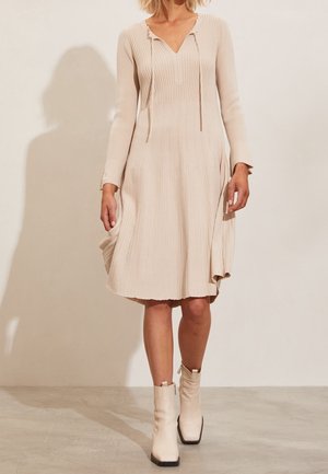 Un mannequin porte une robe midi côtelée couleur crème, à manches longues et à col en V, avec des bottines assorties.