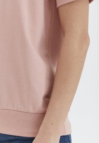Helles Rosa, kurzärmliges Oberteil mit einer weichen Textur und einem Bund. Sichtbarer Arm und Hand, die das Kleidungsstück nahe dem Saum halten.