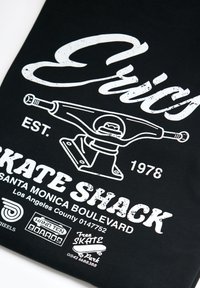 Ilustración de truck de patineta estilo vintage con la marca "Evis", "Skate Shack", Santa Monica Boulevard y logotipos y texto relacionados con el skate en tela negra.