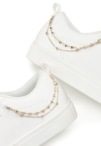 White sneakers made of smooth textile, featuring a layered gold chain accent with heart-shaped details on the heel. Flat rubber sole.