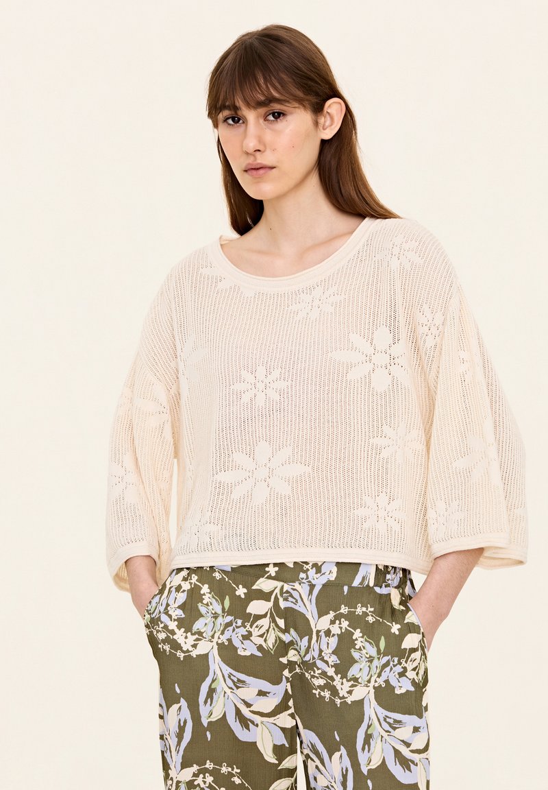 Krämfärgad stickad tröja med blommiga utskärningar, oversized passform, vida ärmar och rundad halsringning. Tillsammans med olivgrön mönstrad byxa.